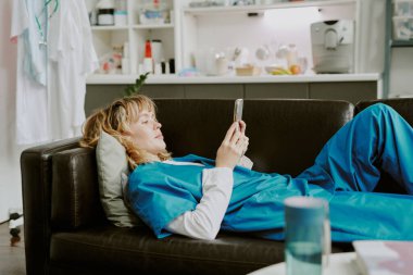 Genç beyaz hemşire, çağrı odasındaki siyah deri koltukta yatarken akıllı telefon kullanıyor.