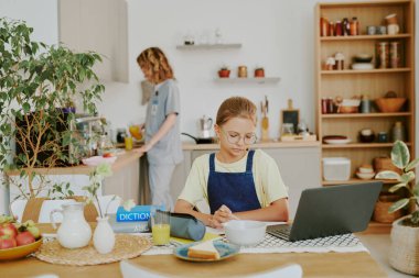Doktor annesi okul ve iş öncesi içecek servisi yaparken beyaz kız masada oturup ödev yapıyor.