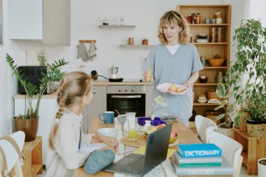Sarışın sağlık görevlisi beyaz anne yemek masasında oturan ve okula hazırlanan kızıyla öğle yemeğini tartışıyor.