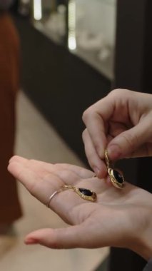 Premium kuyumcuda zarif hediyeler seçen iki etnik kadının dikey yakın çekimi. Palmiyesinde siyah akik desenli altın küpeler tutan kadın.