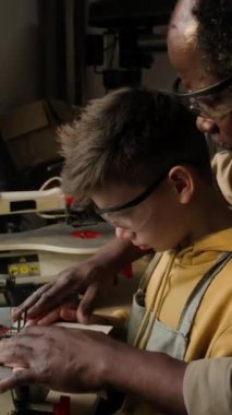 Küçük çocuğun marangozluk atölyesinde marangozluk kursu sırasında marangozluk öğretmeninin yardımıyla testereyle kestiği dikey çekim.