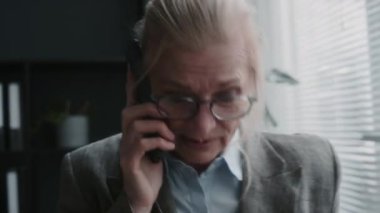 Ofisteki pencerenin yanında duran kıdemli iş kadını telefonla konuşuyor.