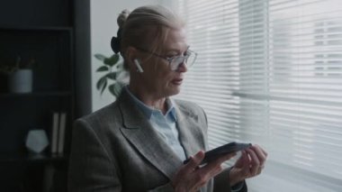 Şık giyinmiş, gözlüklü üst düzey iş kadını pencerenin yanında duruyor ve iş günü kablosuz kulaklıkla konuşuyor.