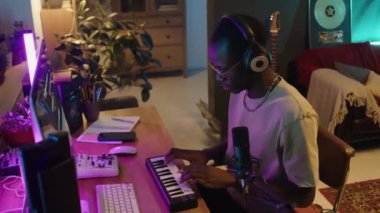African American music producer in headphones using MIDI keyboard in home studio with computer, microphone and guitar in background