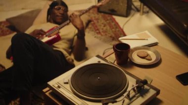 Genç siyah adam evde yerde uzanıyor ve keyfi ile gitar çalıyor, çay fincanı ve aperatifleri yanında.