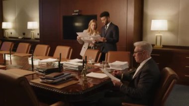Kıdemli gri saçlı avukat toplantı odasında oturuyor ve iki meslektaşı arka planda çalışırken belgeleri inceliyor.