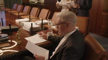 İki asistan ahşap masanın yanında durmuş belgeleri tartışırken, kıdemli gri saçlı erkek hukuk şirketi lideri konferans odasında baş masada oturmuş belgeleri kontrol ediyor.