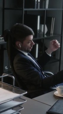 Kablosuz kulaklıklı iş adamının iş yerinde otururken cep telefonuyla konuşması ve dijital tabletteki verileri kontrol etmesi.