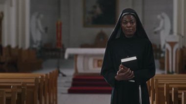 Siyah cüppeli, kutsal kitapla birlikte kilise ayini sırasında kameraya bakan Afrikalı Amerikalı rahibenin portresi.