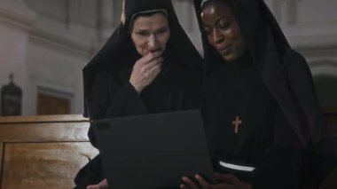 Kilisedeki bankta otururken internetin kablosuz bilgisayarında gezinen iki pozitif etnik rahibenin orta boy fotoğrafı.