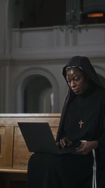 Kilisedeki kablosuz bilgisayarla kilise sıralarında oturan siyah manastırın dikey görüntüsü.