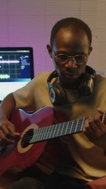 Boynunda kulaklıkları olan, gitar çalan ve kamera önünde gülümseyerek stüdyoda poz veren neşeli siyahi müzisyenin dikey portresi.