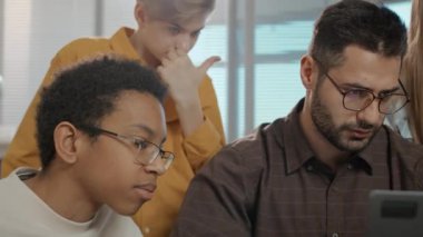 İnternette bilgi arayan dört farklı genç öğrencinin yakın plan fotoğrafları. Genç beyaz erkek öğretmenin rehberliğinde ortak eğitim projesi için dizüstü bilgisayar kullanıyorlar.