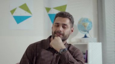 Kahverengi tişörtlü, kendine güvenen beyaz erkek öğretmenin portresi. Sınıfta çene ve çene üzerine oturmuş kameraya bakıyor.