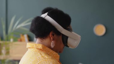 Siyah, yaşlı kadın, sanal gerçeklik kulaklığı takıyor oturma odasında duruyor ve evde yeni aletler denerken etrafa bakıyor.