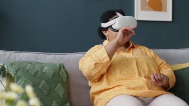Sanal gerçeklik gözlüklü Afrikalı Amerikalı yaşlı kadının evdeki kanepede otururken ve görünmeyen dokunmatik ekranı kullanırken zoom çekimi.