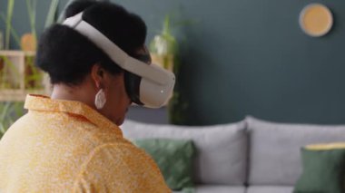 Siyah, son sınıf öğrencisi, sanal gerçeklik gözlüğü takan, oyun oynamayı seçen, elini ileri doğru hareket ettiren ve oturma odasında dururken görünmez dokunmatik ekrandaki düğmeye basan kadın.