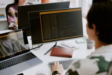 Bilgisayarlarla çevrili erkek programcının, kodlama sırasında başlatılan uygulamayla ekrana bakarken omuz üstü görüntüsü, iş arkadaşı önünde oturuyor.