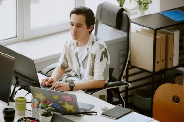 Bilgisayarlar ve termal kahve bardaklarıyla çevrili rahat bir ofiste çalışırken klavyede yazan ve bilgisayar ekranına bakan genç bilişim uzmanı.