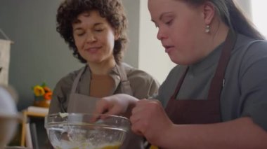 Önlüklü, mutfak masasında oturan ve mutfak kursu sırasında kadın öğretmenin kontrolü altında cam kasede yemek malzemeleri karıştıran bir kız.