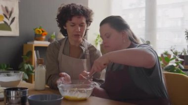 Kadın öğretmen cam kaseye süt eklerken Down sendromlu kız yemek pişirme dersinde malzemeleri karıştırıyor.