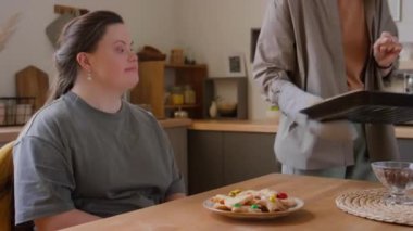 Down sendromlu bir kız mutfak masasında otururken anne evde gün boyunca tabakta pişmiş kurabiye servis ediyor.