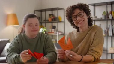 Down sendromu olan neşeli bayan öğretmen ve kızın portresi origami tilki suratları taşıyor ve oturma odasında birlikte otururken kameraya gülümseyerek poz veriyor.