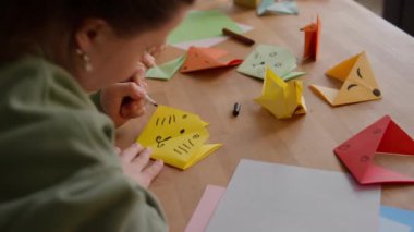 Down sendromlu yaratıcı kızın, kadın öğretmenle sanat dersinde yaptığı origami tilkisine şirin bir yüz çizerken yakın plan çekimi.