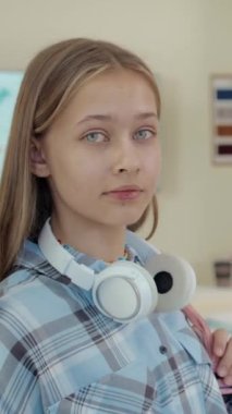 Mavi gözlü genç kızın dikey portresi ortaokul sınıfında duruyor gülümsüyor ve kendinden emin bir şekilde kameraya bakıyor.