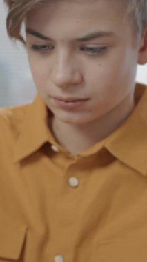 Sarı tişörtlü genç bir çocuğun dikey portresi sınıfta oturmuş akıllı telefon kullanıyor ve neşeyle gülümsüyor.