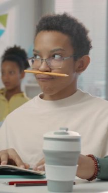 Gözlüklü genç Afrikalı Amerikalı bir çocuğun dikey portresi. Ders sırasında okulda masasında oturup dudak ve burun arasına kalem koyarak maskaralık yapıyor.