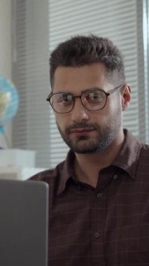Genç, kendine güvenen beyaz erkek öğretmenin dikey portresi ya da okulda masasında oturan gözlüklü, gülümseyen ve dizüstü bilgisayarla çalışırken kameraya bakan müdür.