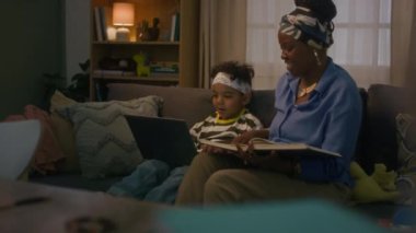 Orta uzunlukta bir Afrikalı Amerikalı kadının oğluna kitap okurken kanepedeki kablosuz bilgisayarı kullanırken görüntüsü.