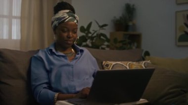 Evdeki kanepede kablosuz dizüstü bilgisayar üzerinde çalışırken kameraya gülümseyen Afro-Amerikan kökenli neşeli kadın solofenatörün orta boy portresi.
