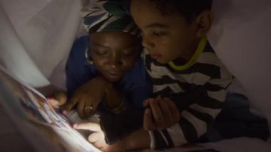 Gece battaniyenin altında yatarken annesine fener okuyup çizgi roman okuyan Afrikalı Amerikalı çocuğun orta boy fotoğrafı.