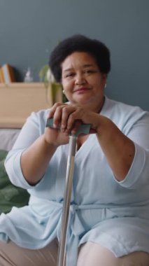 Afro-Amerikan yaşlı bayanın dikey portresi. Kameraya bakıyor ve bastonunun önünde otururken el ele tutuşuyor.