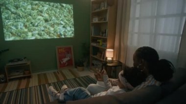 Oturma odasındaki duvara yansıtılan böceklerle ilgili film izlerken siyah kadının oğluna sarıldığı arka planda.