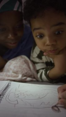 Gece geç saatlerde oğlunun yanında el feneri tutan ve kağıda resim çizen Afro-Amerikan annenin dikey anahtarı.