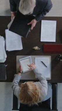 Hukuk firmasında belgelerle çalışan iki profesyonel avukatın dikey aşağı bakışı, adamın evrak klasörünü masaya koyması, kadının dijital tablette metin dosyasını kaydırması