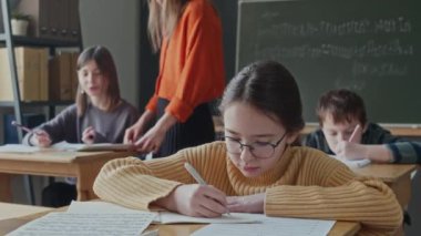 İlkokulda masasında otururken müzik dersi alan küçük bir kız. Öğretmen arka planda başka bir öğrenciyle konuşuyor.