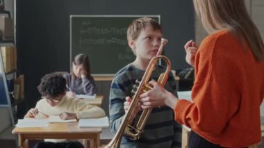 İlkokul öğrencisi, müzik dersi sırasında yanında trompetle duran bayan öğretmeninin açıklamasını dinliyor. Sınıf arkadaşları arka planda yazım sınavı yapıyor.