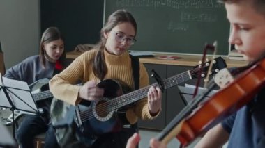 Gitar çalan iki kız ve müzik dersinde keman çalan bir oğlan.