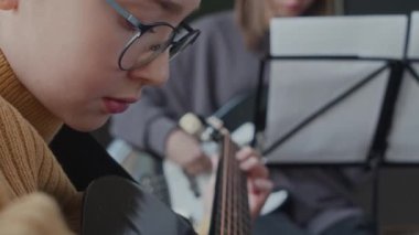 Okuldaki müzik sınıfında akustik gitarla şarkı söyleyen genç bir kızın yakın plan çekimi, arka planda enstrüman çalan bir sınıf arkadaşı.