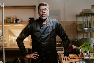 Çok deneyimli siyah üniformalı bir şef mutfak tezgahının yanında duruyor ve doğrudan kameraya bakıyor.