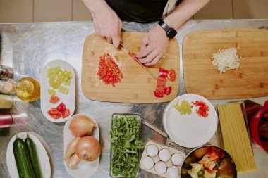 Üst görüntüde tanınmayan erkek elleri, çelik mutfak tezgahında sebzelerle birlikte bulunan ahşap kesme tahtasıyla sebze kesiyor.