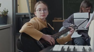 Müzik dersi sırasında akustik gitarıyla otururken kameraya bakan okul çocuğunun portresi, arka planda enstrüman çalan kız