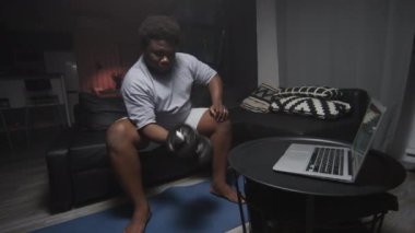 Obeziteli genç bir adam oturma odasında kanepede oturuyor, halterli kıvrımlar yapıyor ve evde yoğun bir egzersizden sonra yoruluyor.
