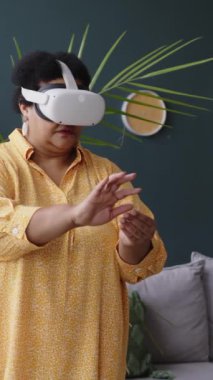 Siyah yaşlı bir kadının sanal gerçeklik gözlüğü takıp oturma odasında dururken yeni bir aleti nasıl kullanacağını öğrenmesine dikey bakış açısı.