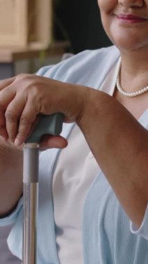 Gülümseyen son sınıf siyah bir kadının dikey portresi. Kolye takıyor, evde oturuyor ve elini bastonun üzerinde tutuyor.