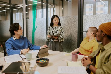 Dijital tableti elinde tutan genç Asyalı yönetici, çok ırklı meslektaşlarının önünde toplantı sırasında proje hakkında konuşuyor.